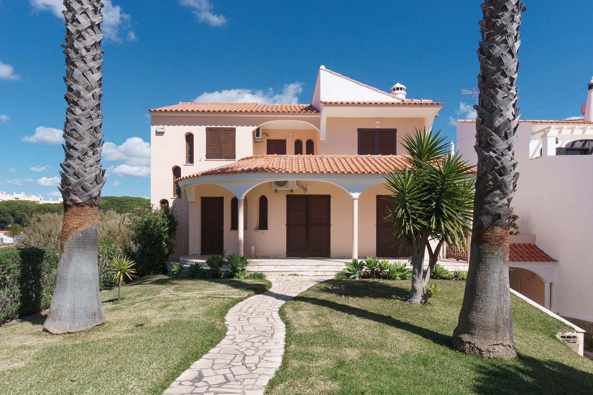 A two-story house with a tiled roof, a stone pathway leading to the entrance, and palm trees in the garden.