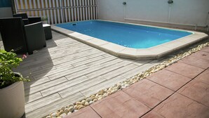 Una piscina rectangular con borde de azulejos, rodeada por una terraza de madera y un borde de piedra.