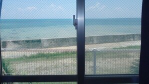 Vista a través de una ventana con celosía metálica, sobre una playa con un muro de hormigón y vegetación.