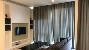 A modern living room with a flat-screen TV mounted on a white wall, a black glass coffee table, a grey sofa, and grey curtains. - Best Homestay (Kuala Lumpur)