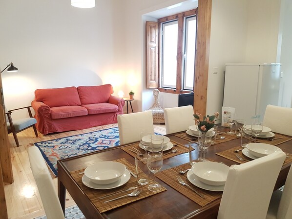 A dining area with a wooden table set for four, surrounded by white chairs. A red sofa and a small side table with a lamp are visible in the background. - Charming & Central Apartment in Traditional Chalet (Sintra)