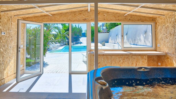 A modern spa room with a hot tub, large glass doors leading to an outdoor pool area, and a view of the surrounding greenery. - Villa Emanuele Costarainera: wonderful modern villa, for 16 ppl, with garden, heated pool, hot-tub (Costarainera)