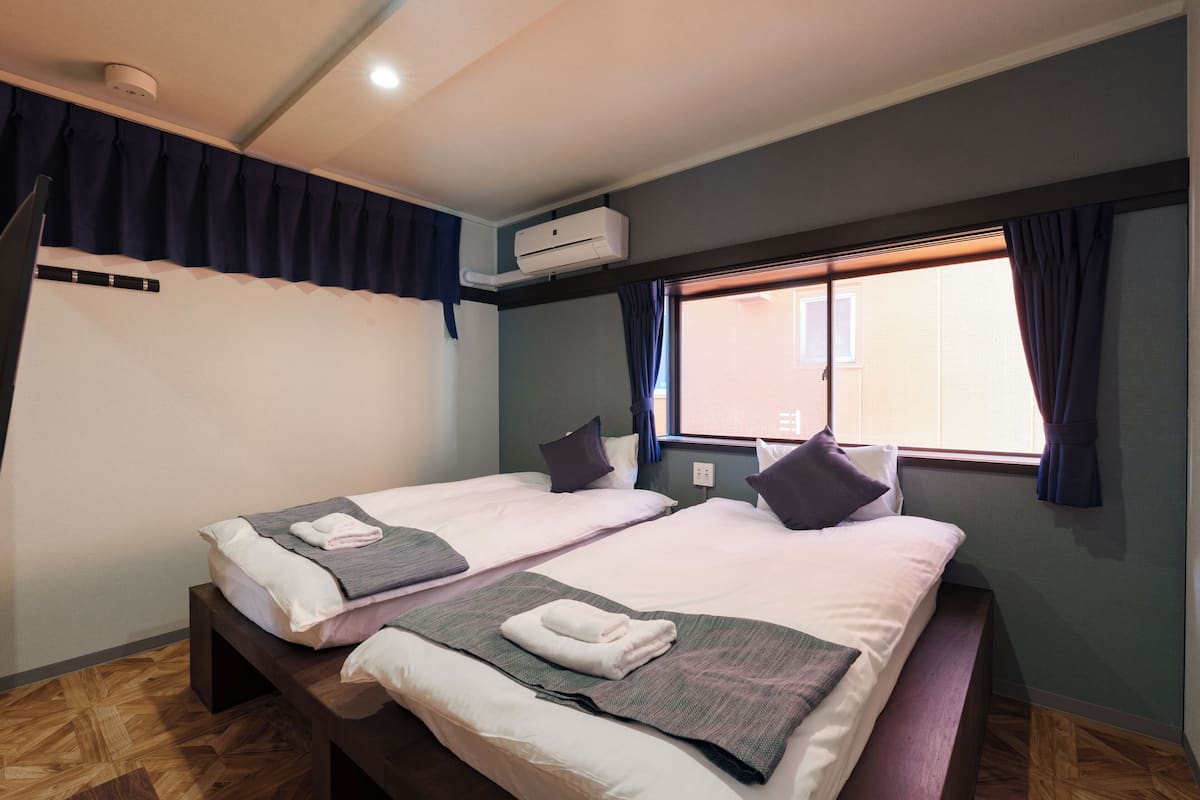 A hotel room with two beds, a window with curtains, an air conditioning unit, and a wooden floor.