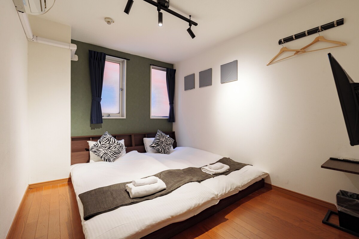 A neatly made bed with pillows, a bedside table, a wall-mounted TV, and a window with curtains.