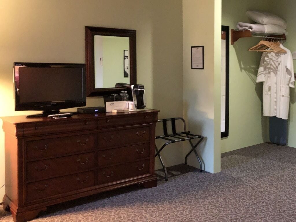 A room with a wooden dresser, a television, a mirror, a coat rack with a white robe, and a framed certificate on the wall.