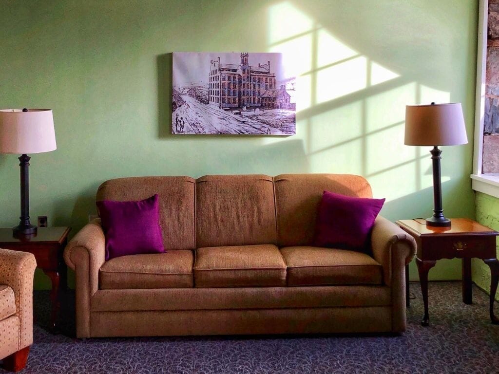 A room with a brown sofa, two purple cushions, a painting on the wall, and two lamps on side tables.