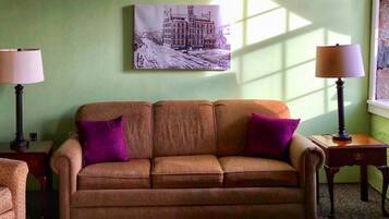 A room with a brown sofa, two purple cushions, a painting on the wall, and two lamps on side tables.
