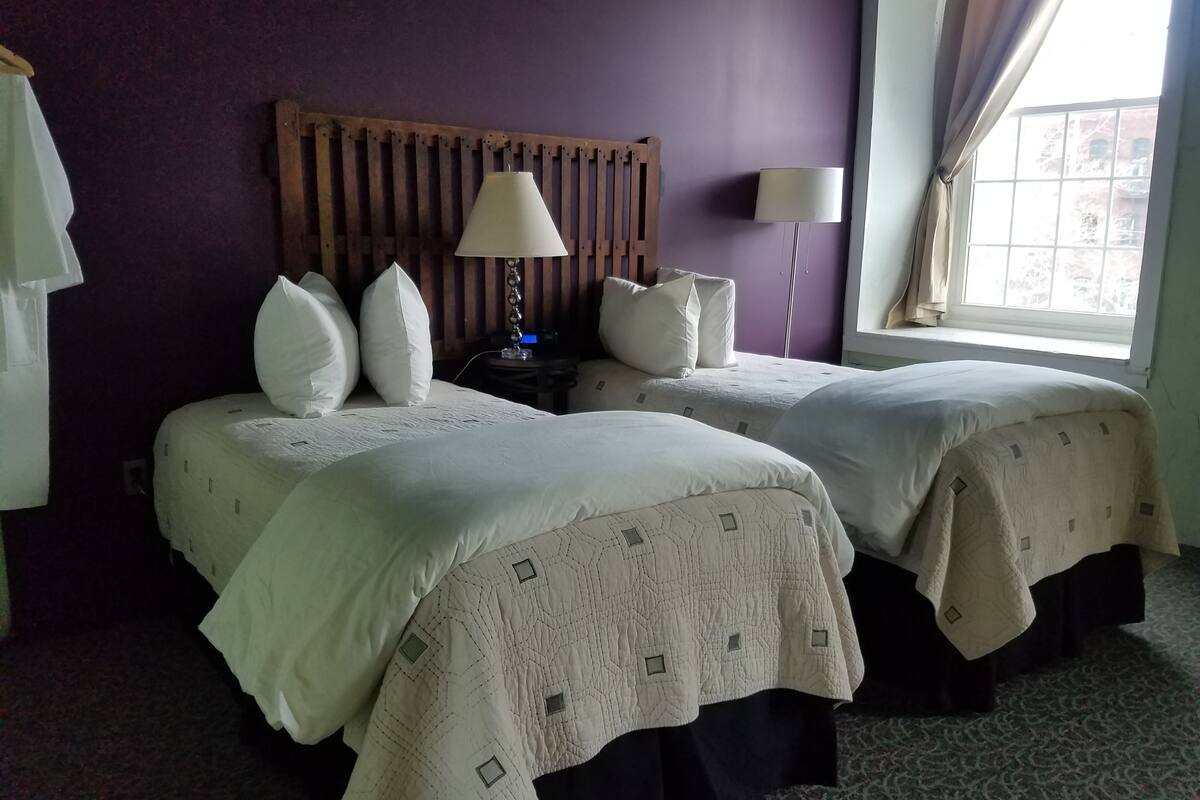 A hotel room with two beds, a wooden headboard, a nightstand with a lamp, and a window with curtains.