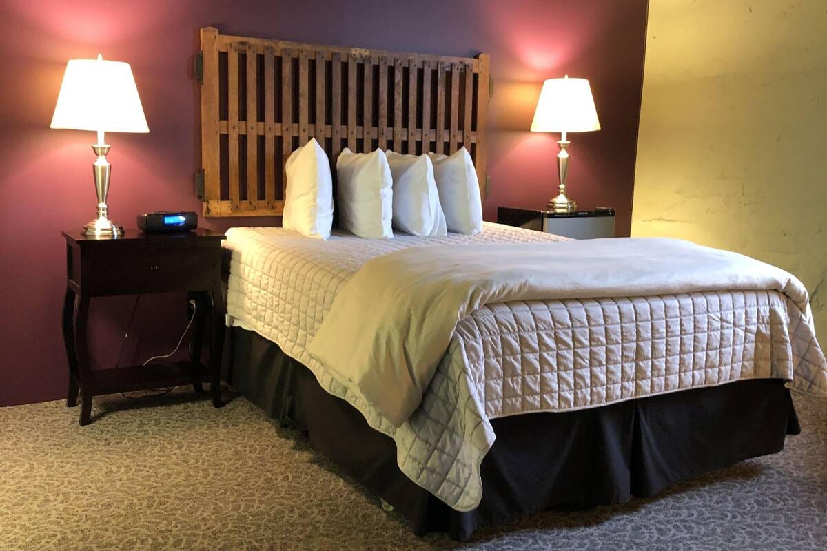 A neatly made bed with a quilted bedspread and a wooden headboard. There are two bedside tables with lamps, and a small nightstand with a clock. The room has a carpeted floor and a plain wall.