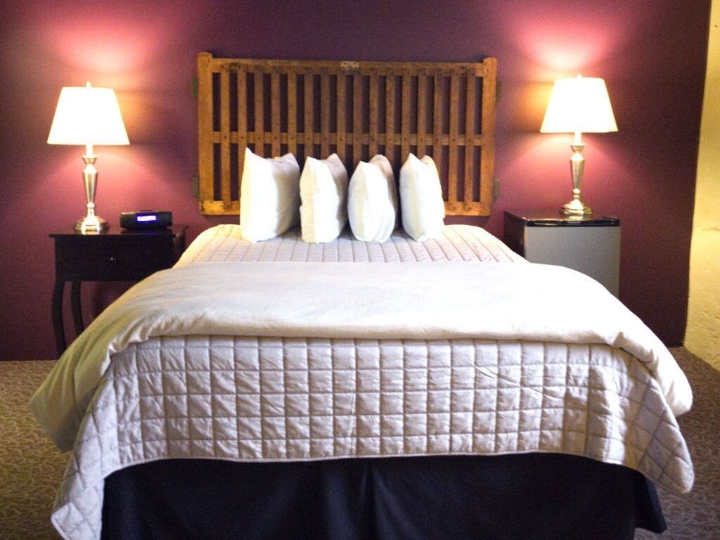 Una cama bien hecha con cubrecama acolchado, flanqueada por dos mesitas de noche con lámparas, y un cabecero de madera.