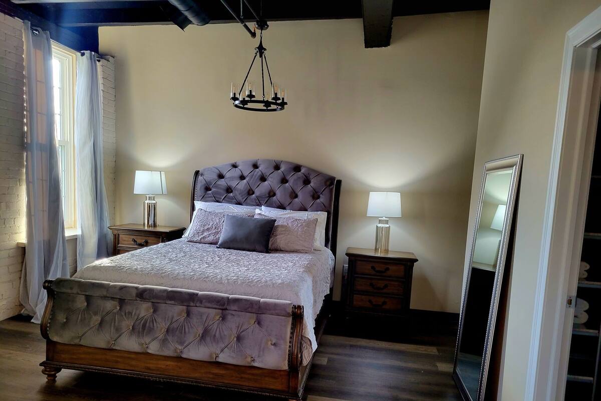 A bedroom with a large bed, two bedside tables with lamps, a hanging chandelier, and a mirror on the wall.