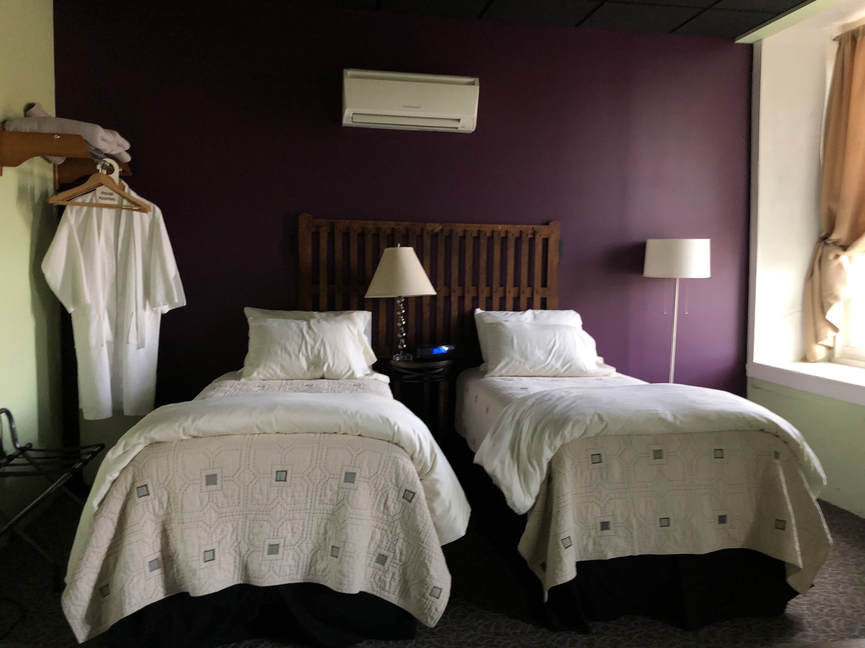 Una habitación con dos camas, un perchero, una mesita de noche con lámpara y una ventana con cortinas.