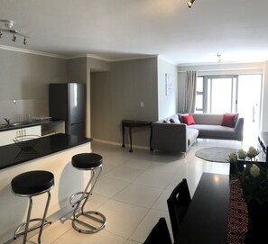 A modern kitchen with a refrigerator, a dining area with a table and chairs, a sofa, and a large window. - 606 The Beaumont (Cape Town)