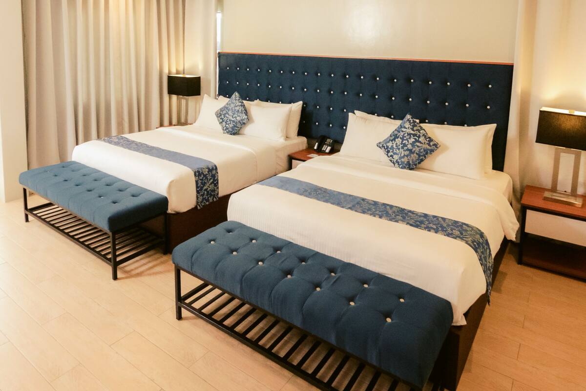 Une chambre d’hôtel avec deux lits, un banc, une lampe et un rideau.