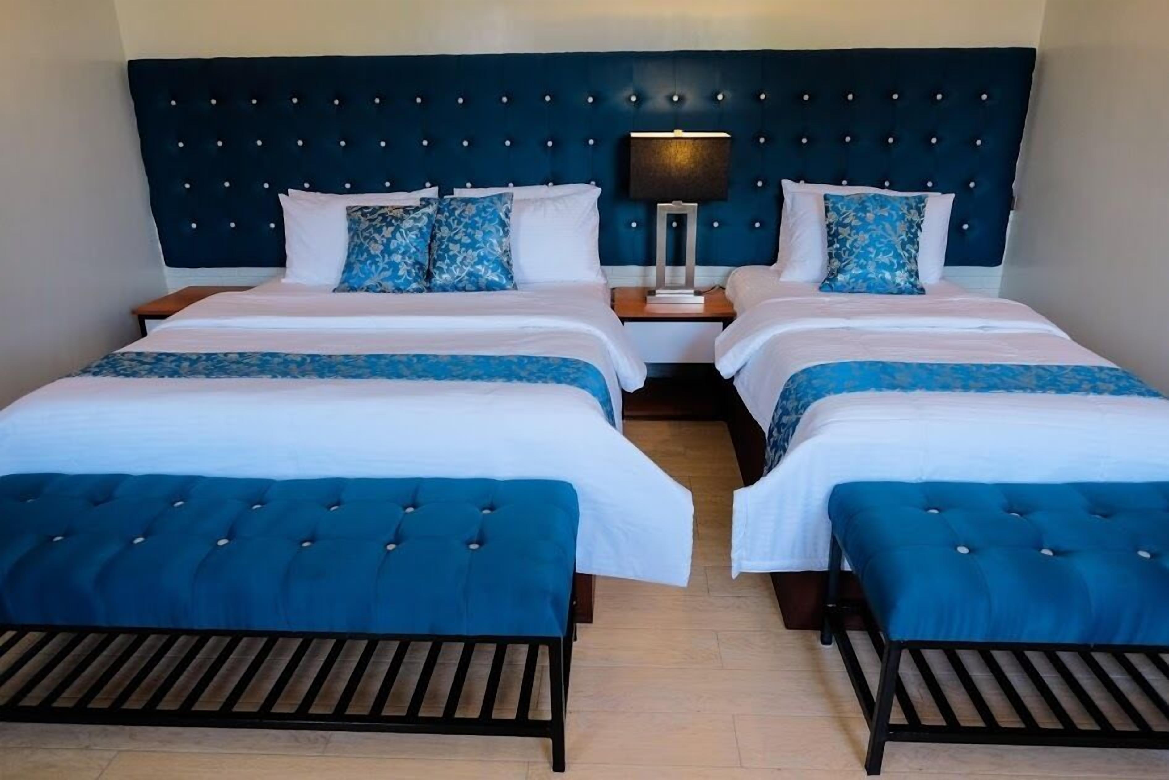 A hotel room with two beds, each with a blue upholstered headboard and matching footboard. There are two bedside tables with lamps, and the room has a dark blue headboard with a tufted design.