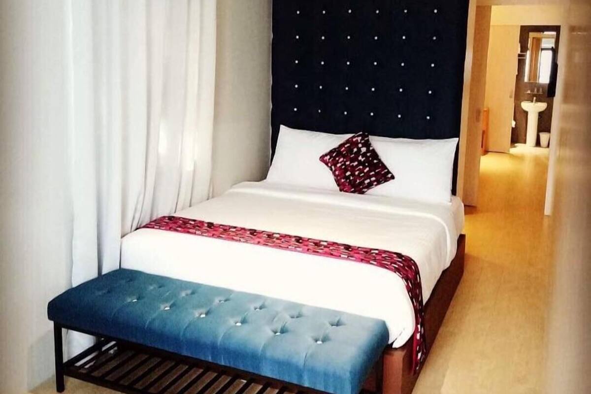 Une chambre d’hôtel avec un grand lit, une banquette, une tête de lit sombre et une vue sur la salle de bain.