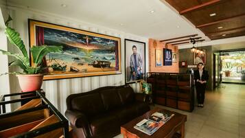 A lobby with a leather sofa, a coffee table with magazines, a large painting of a beach scene, and a reception desk with a person standing behind it.