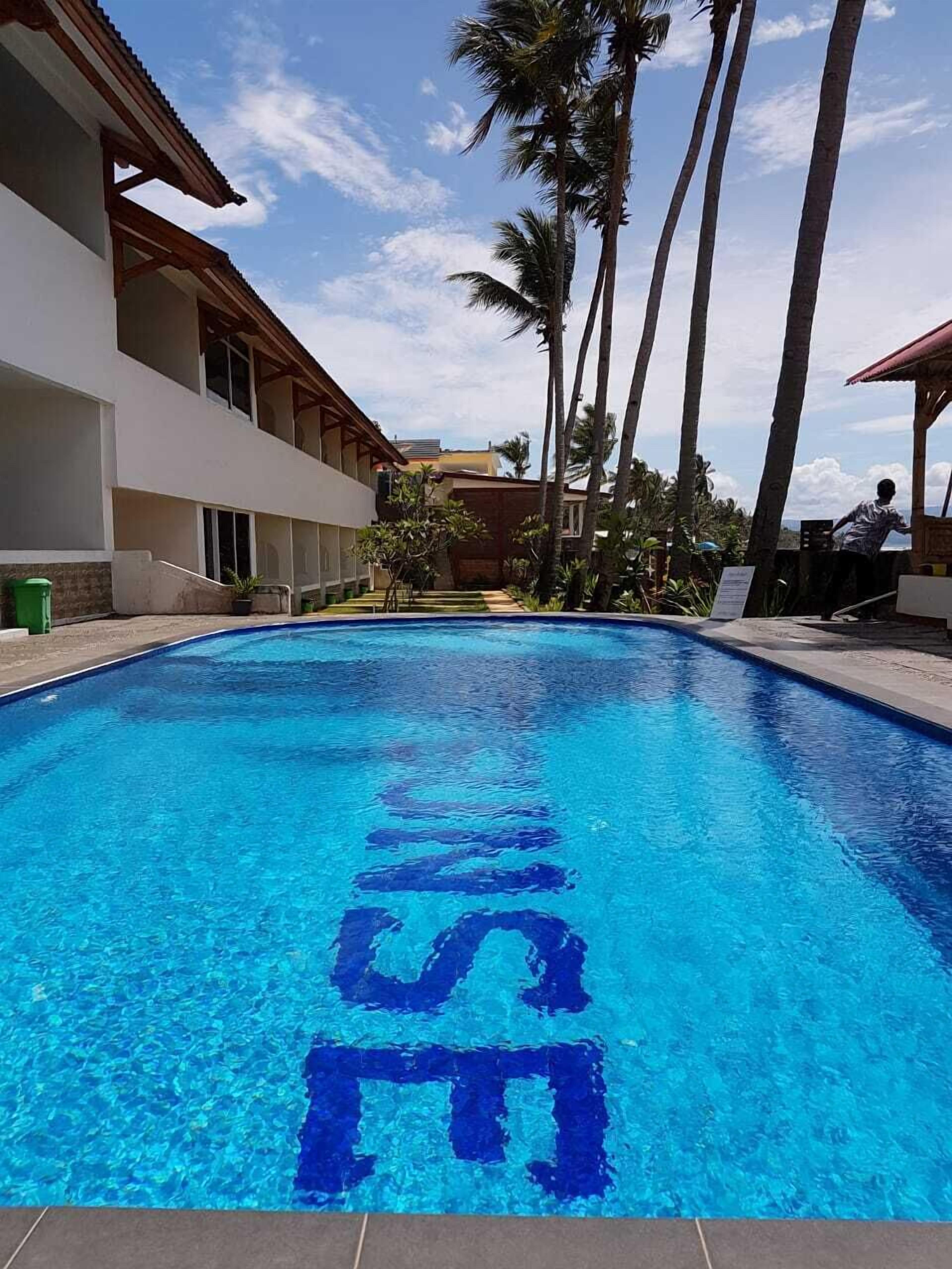 Une piscine à l’eau d’un bleu limpide, entourée de palmiers et bordée d’un bâtiment doté d’un balcon.