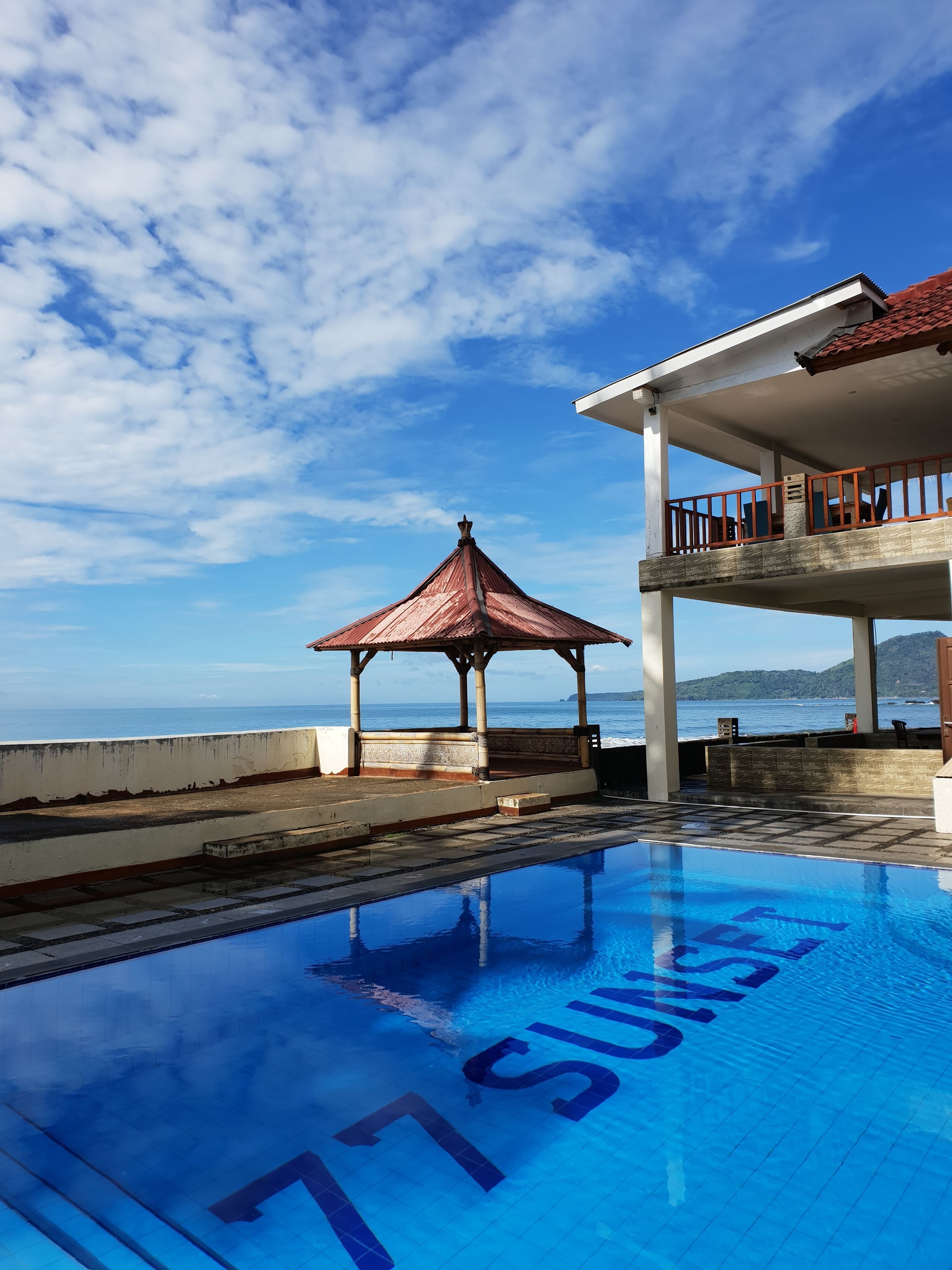 A swimming pool with the words "77 SUISEN" on the surface, a gazebo, and a building with a balcony overlooking the sea.
