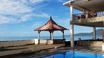 A swimming pool with the words "77 SUISEN" on the surface, a gazebo, and a building with a balcony overlooking the sea.