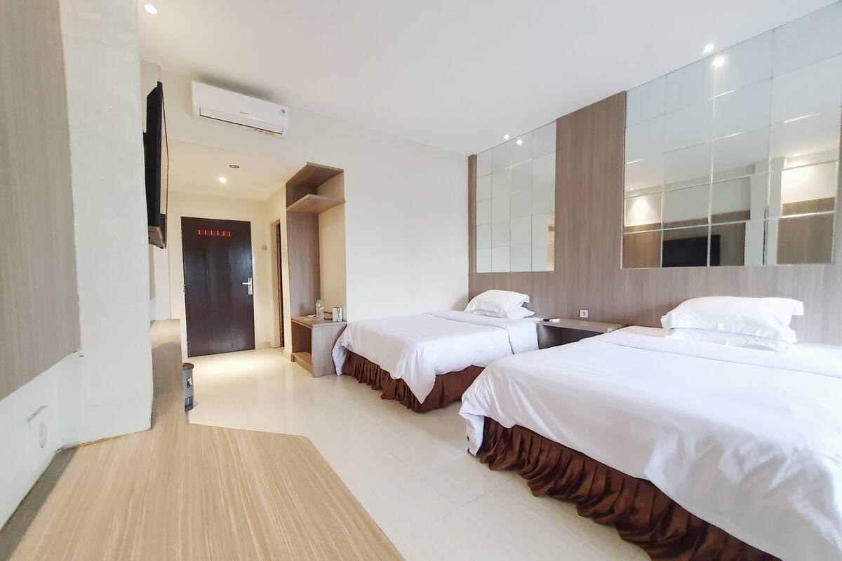 A hotel room with two beds, a wooden floor, a large mirror, and a door leading to another room.