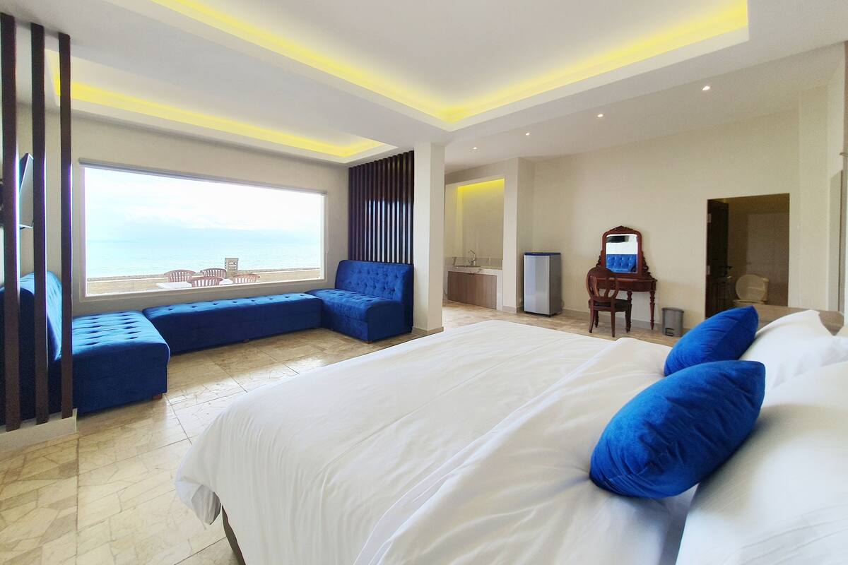 A modern hotel room with a large bed, a seating area by the window, and a view of the sea.