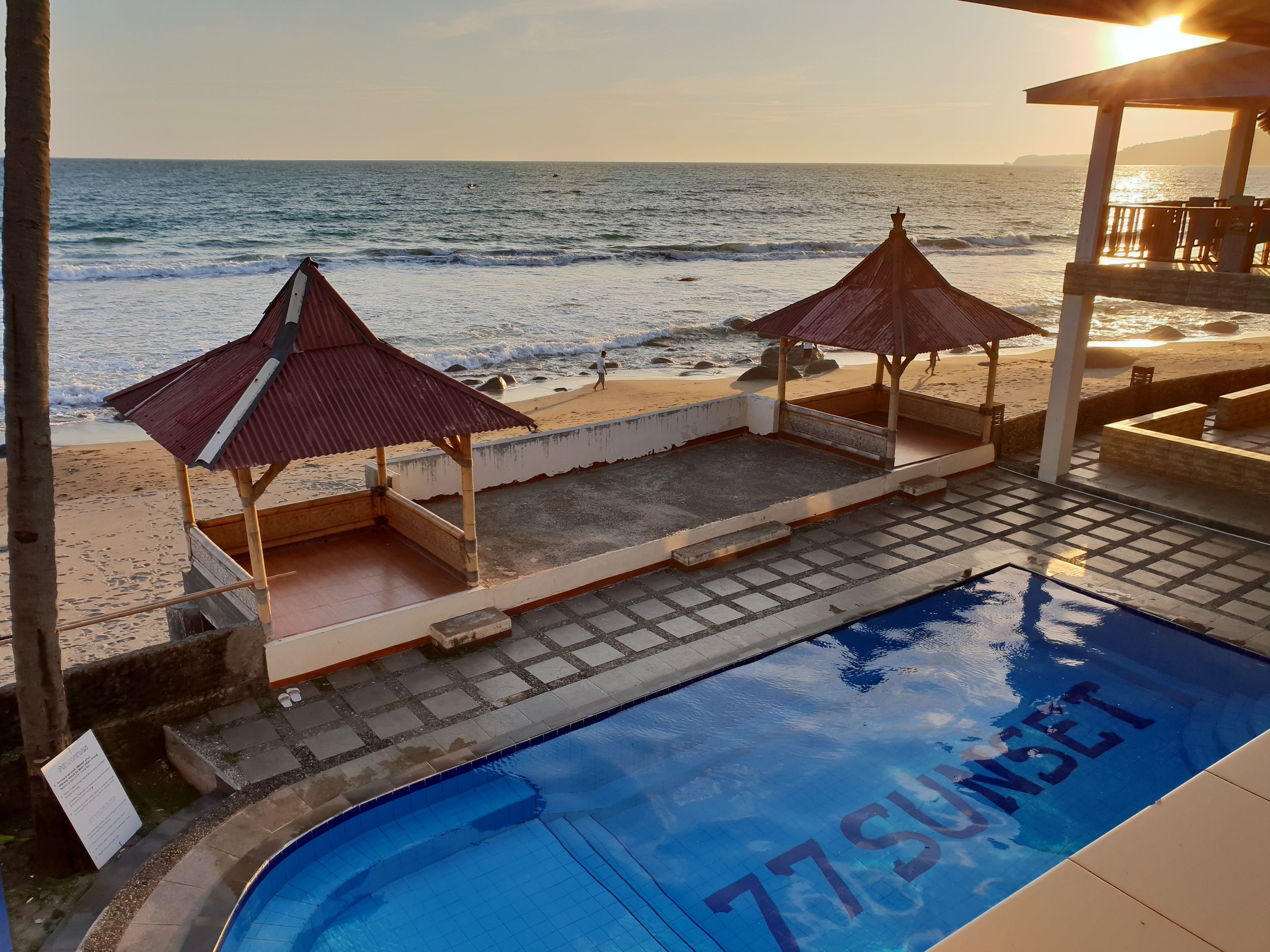 Une piscine avec l’inscription « 77SUNSET » sur le bord, deux parasols de plage et une vue sur la plage.
