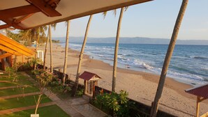 A beach view from a covered area with palm trees, a sandy beach, and a small structure. - 77 Sunset Plaza (Cisolok)