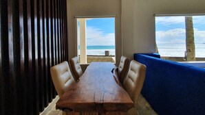 A dining area with a wooden table and chairs, a blue cushioned bench, and a view of the ocean through large windows. - 77 Sunset Plaza (Cisolok)