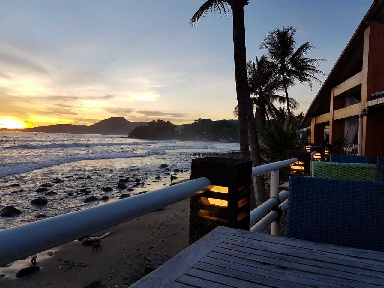 Vista da praia com um deque de madeira, palmeiras e um prédio com varanda.