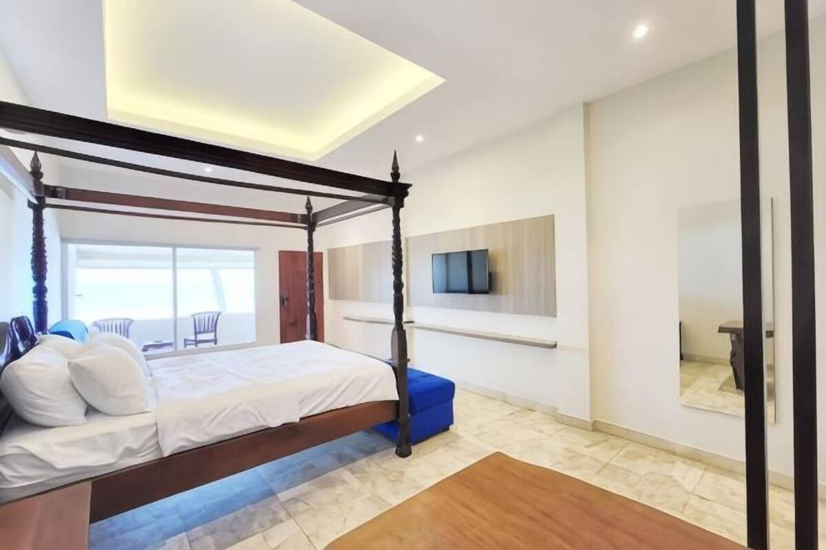 A four-poster bed with a wooden frame, a flat-screen TV mounted on the wall, and a large window offering a view of the outdoors.
