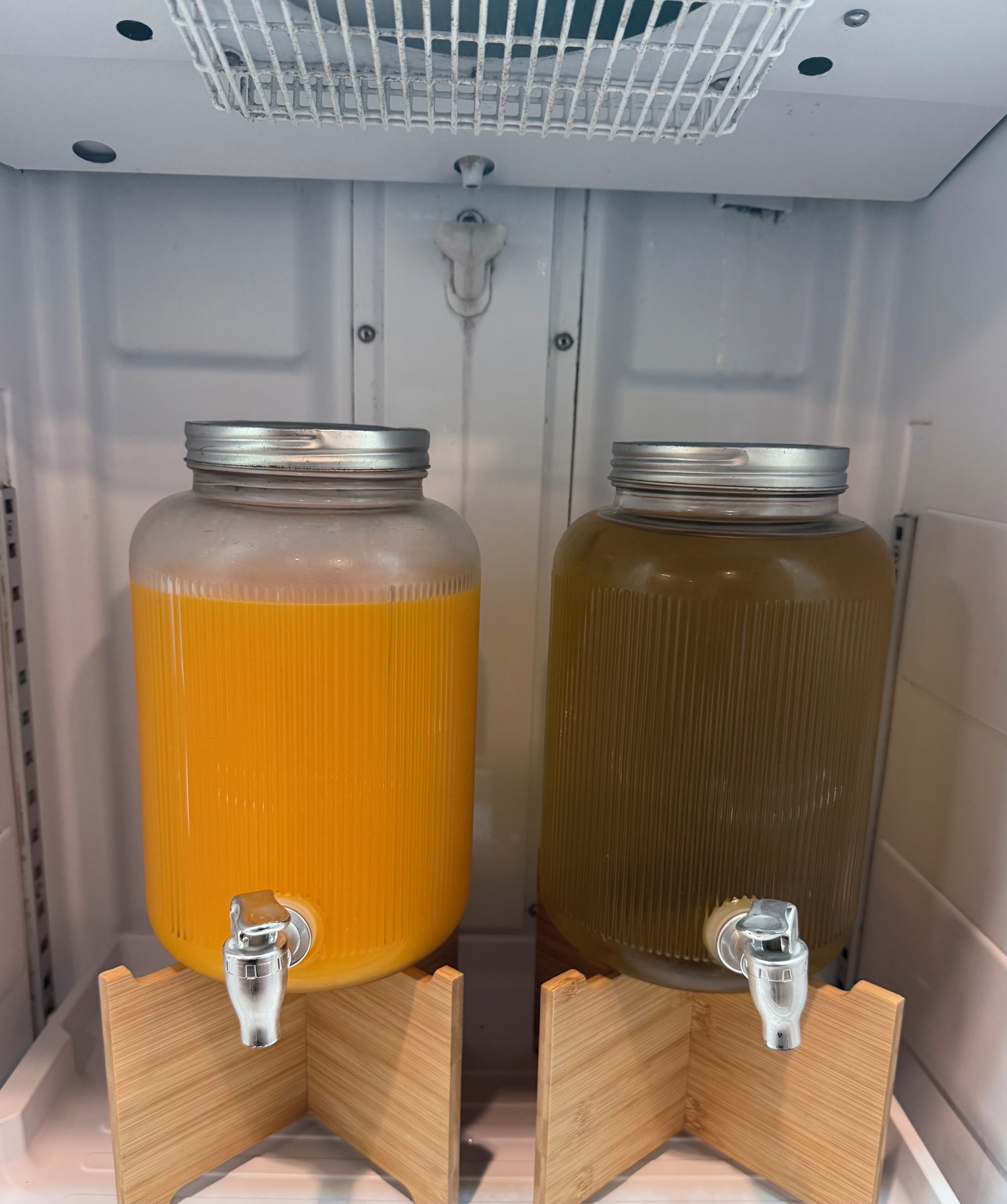 Deux grands bocaux en verre dotés de couvercles métalliques, chacun muni d’un robinet fixé à un support en bois, placés à l’intérieur d’un réfrigérateur.