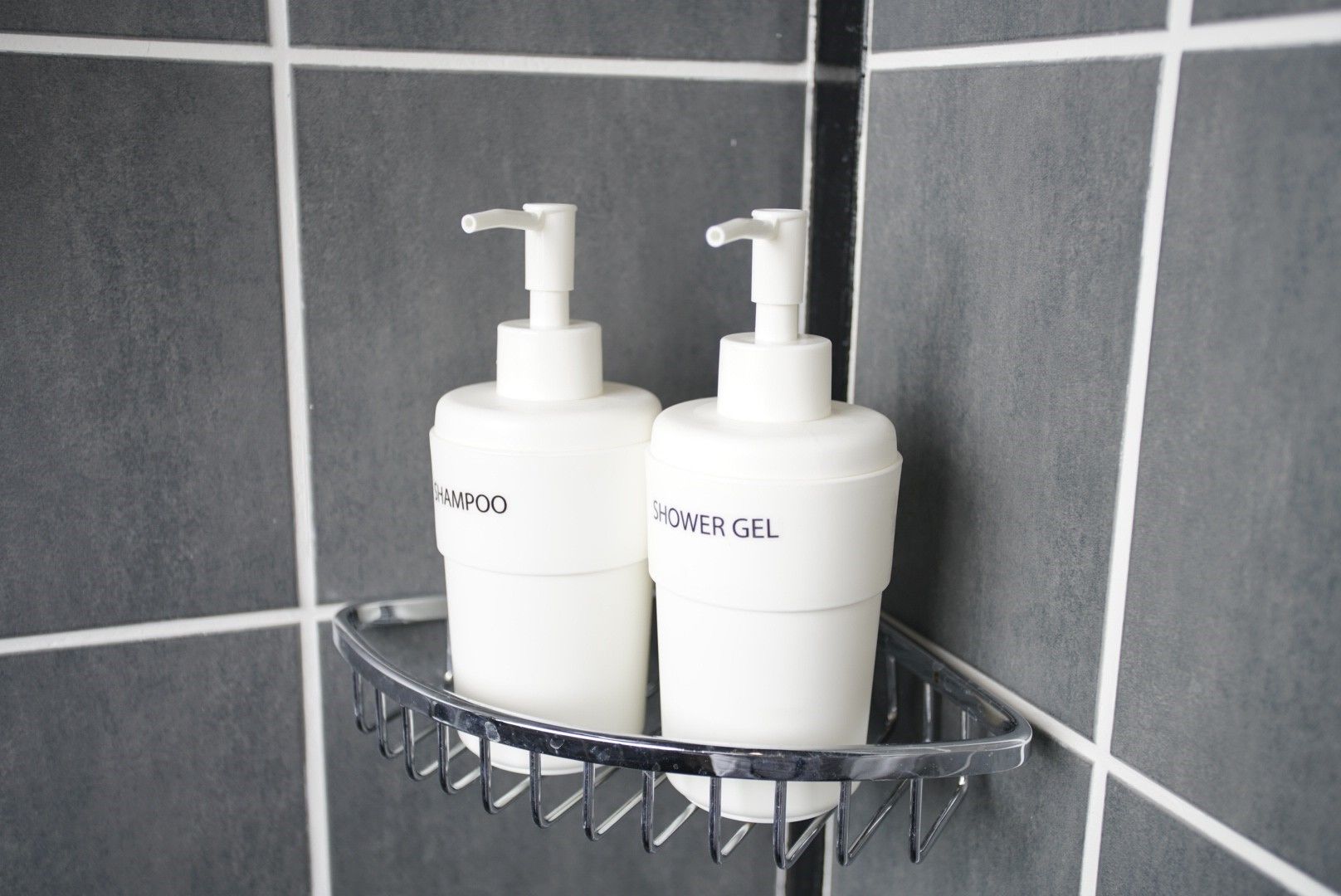 To hvide flasker med teksten 'SHAMPOO' og 'SHOWER GEL' står på en metalreol foran en flisevæg.