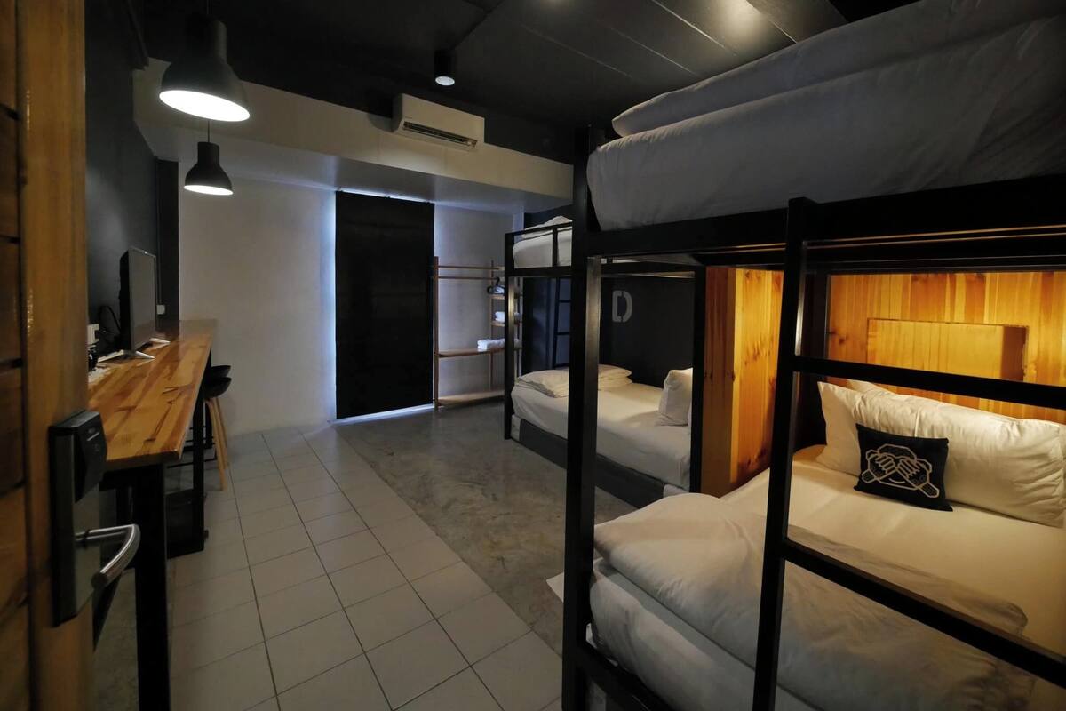 A bunk bed room with a wooden desk and a computer, a hanging light fixture, and a door leading to another room.