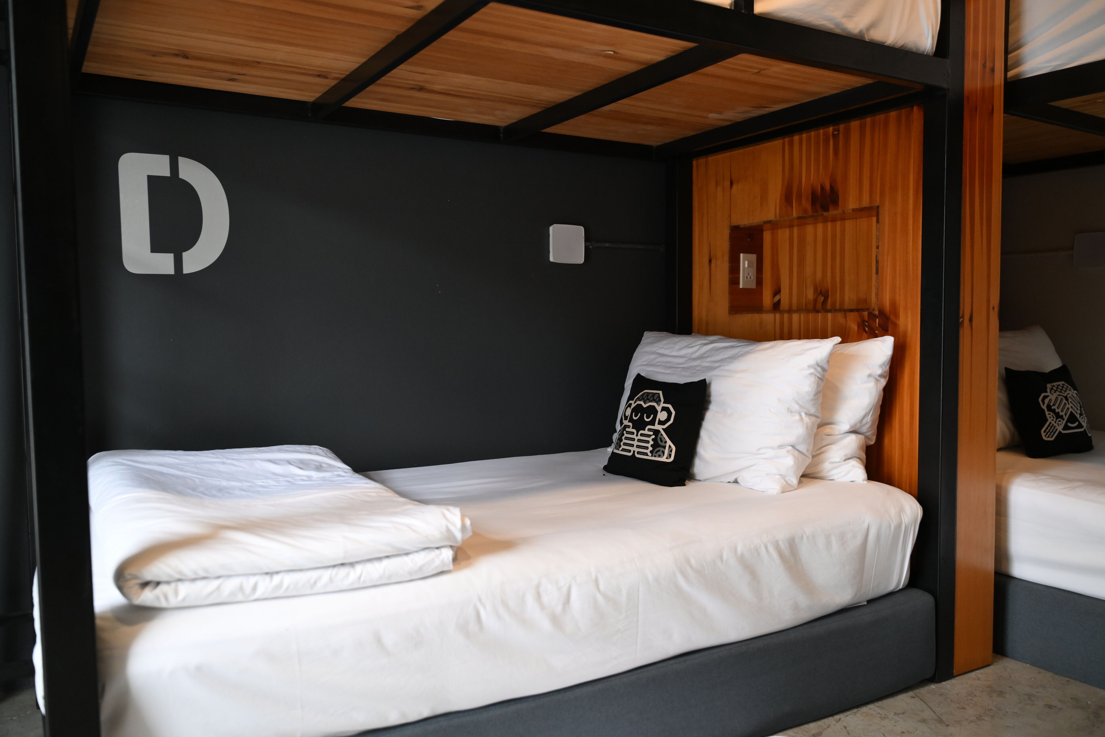 Ein Stockbett mit weißer Bettwäsche, eine schwarze Wand mit dem weißen Buchstaben 'D' und eine Holzzielende.