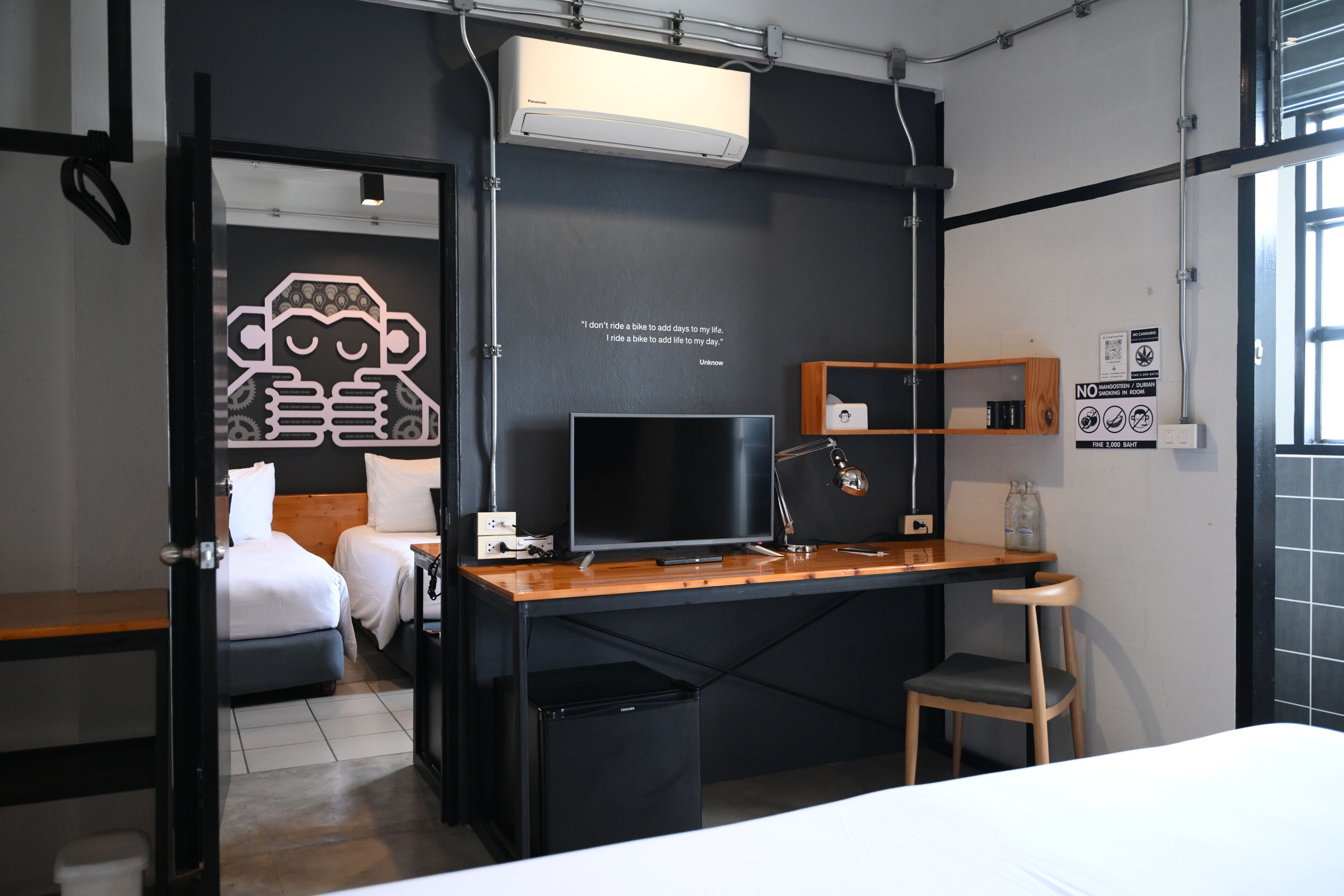 Ein modernes Hotelzimmer mit Bett, Schreibtisch, Stuhl, Fernseher und einer Wand mit einem Zitat.