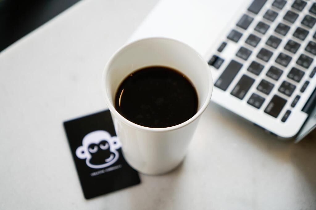 A white cup of coffee on a table with a laptop and a black card with a white design.