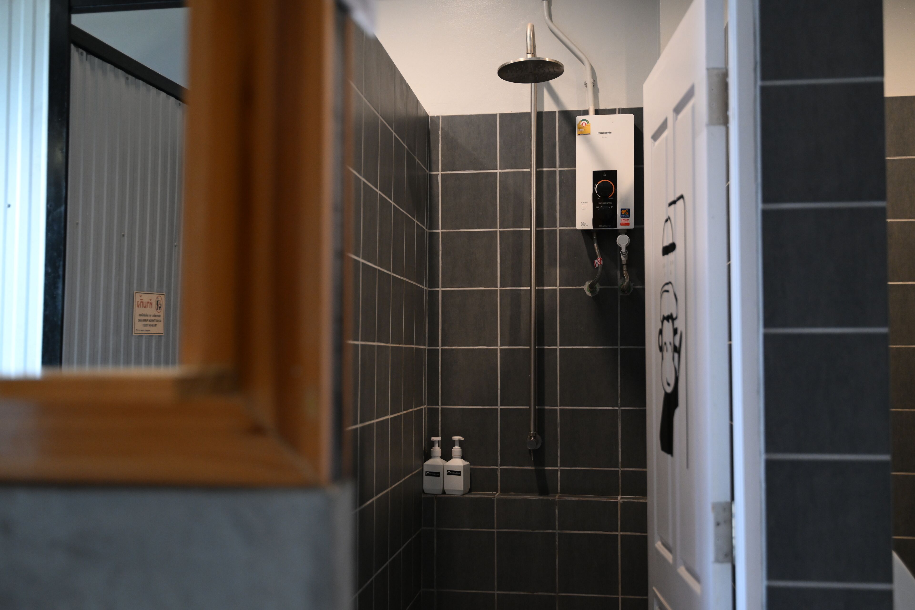 A bathroom with dark tiled walls, a shower with a handheld showerhead, and two bottles of shampoo on the floor.