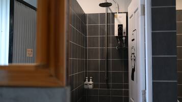 A bathroom with dark tiled walls, a shower with a handheld showerhead, and two bottles of shampoo on the floor.