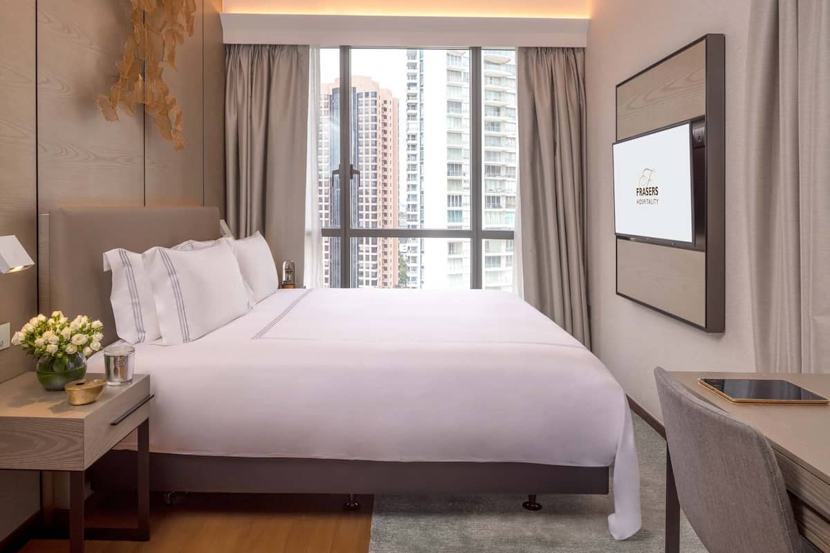 A modern hotel room with a large bed, a desk, a chair, a TV, and a view of tall buildings.