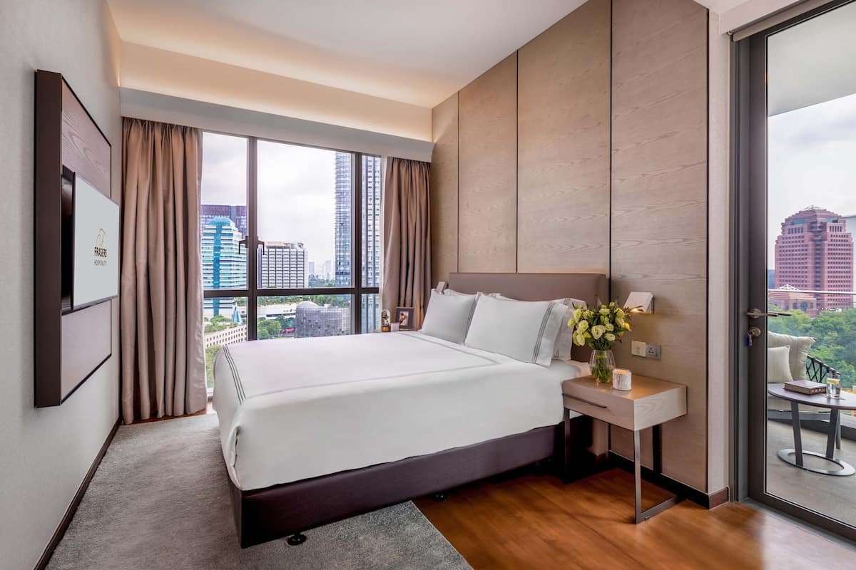 A modern hotel room with a large bed, a bedside table with a vase of flowers, and a city view through large windows.