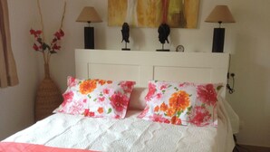 A neatly made bed with a white quilt and pink accents, a vase with red flowers, and a painting of two figures on the wall.