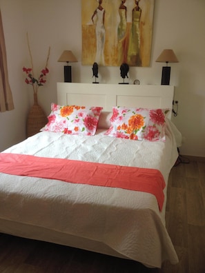 Ein ordentlich bezogenes Bett mit weißer Decke und rosa Akzenten, eine Vase mit roten Blumen und ein Bild von zwei Personen an der Wand.