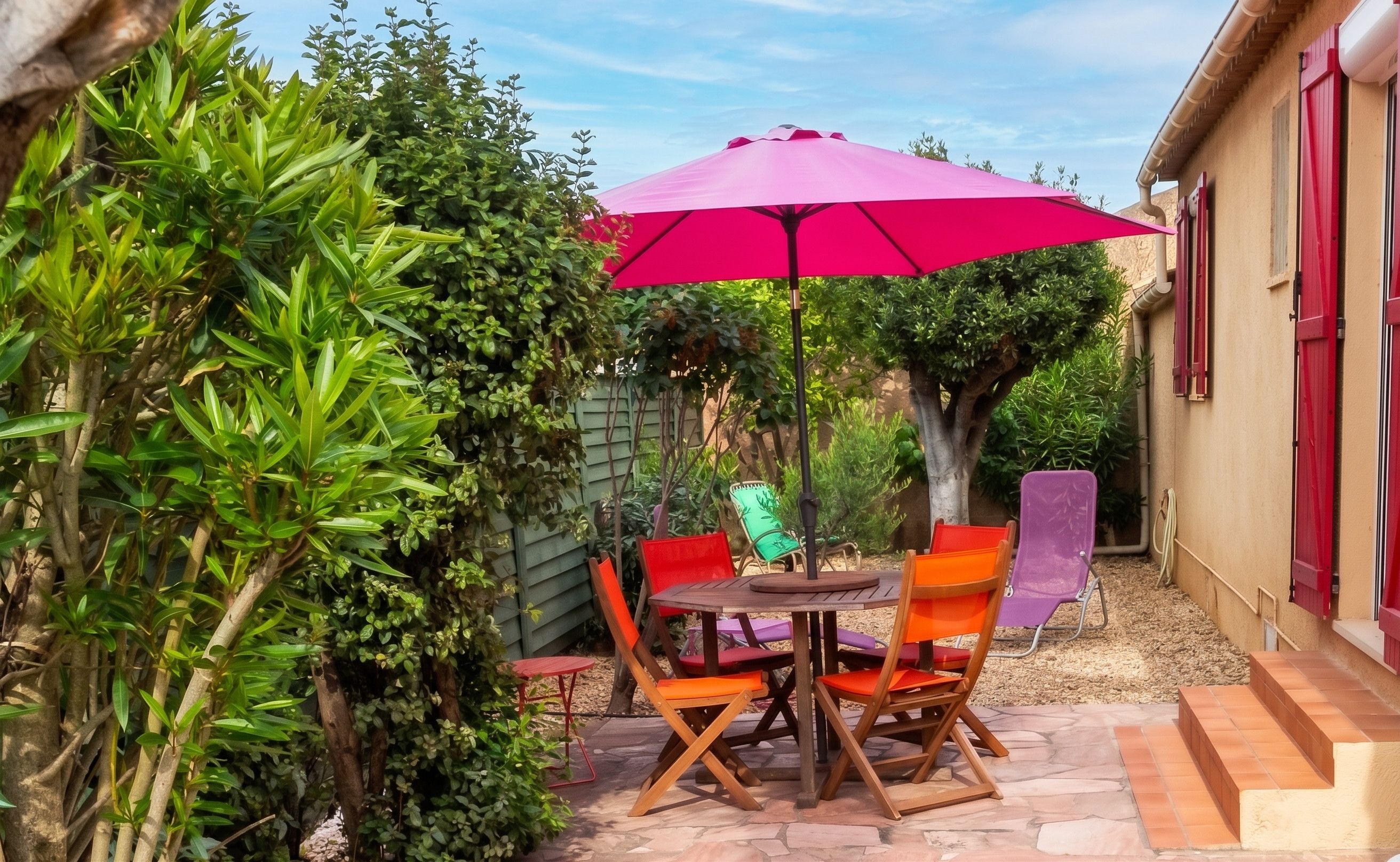Un patio con una mesa, sillas, un paraguas rosa y diferentes plantas.
