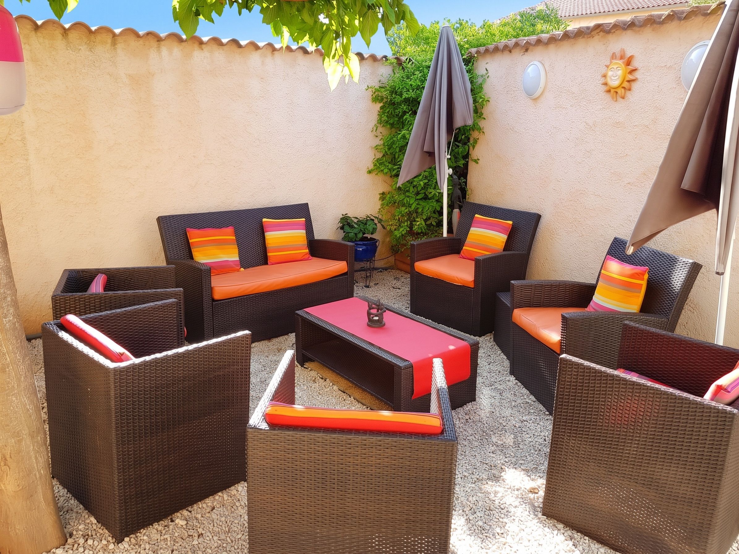 Un espace patio aménagé avec des meubles en osier, comprenant deux canapés et deux fauteuils, chacun agrémenté de coussins colorés. On y trouve une petite table avec un plateau rouge et un élément décoratif en métal. Le patio est pavé de petits galets, et on y aperçoit un pot de fleurs ainsi qu’une décoration murale en forme de soleil.