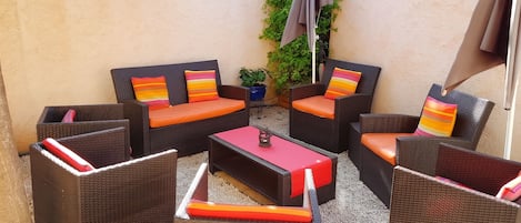 Un espace patio aménagé avec des meubles en osier, comprenant deux canapés et deux fauteuils, chacun agrémenté de coussins colorés. On y trouve une petite table avec un plateau rouge et un élément décoratif en métal. Le patio est pavé de petits galets, et on y aperçoit un pot de fleurs ainsi qu’une décoration murale en forme de soleil.