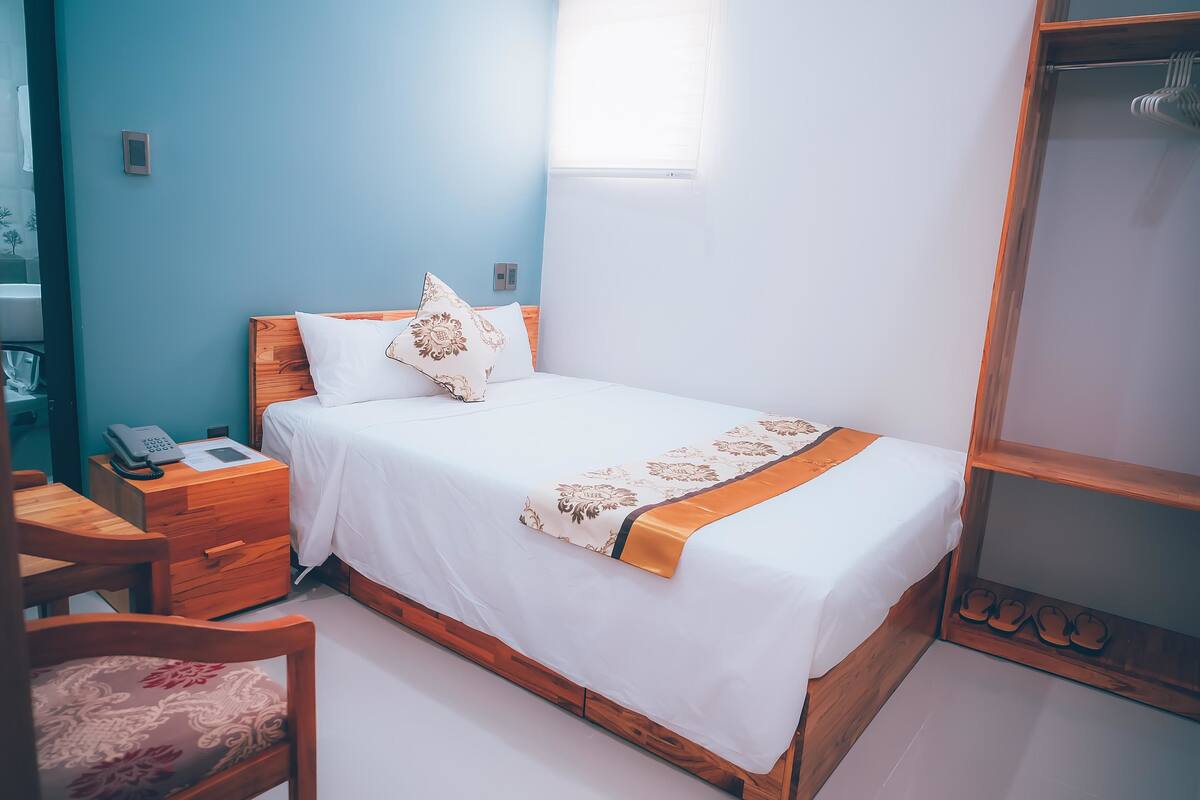A neatly made bed with a wooden headboard, a bedside table with a phone, and a chair with a floral cushion.
