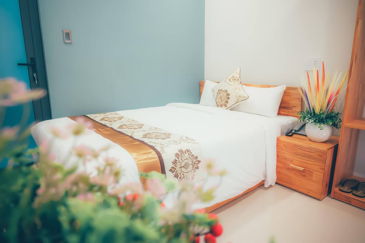 A neatly made bed with a white and gold bedspread, a wooden nightstand with a vase of dried flowers, and a wooden headboard.