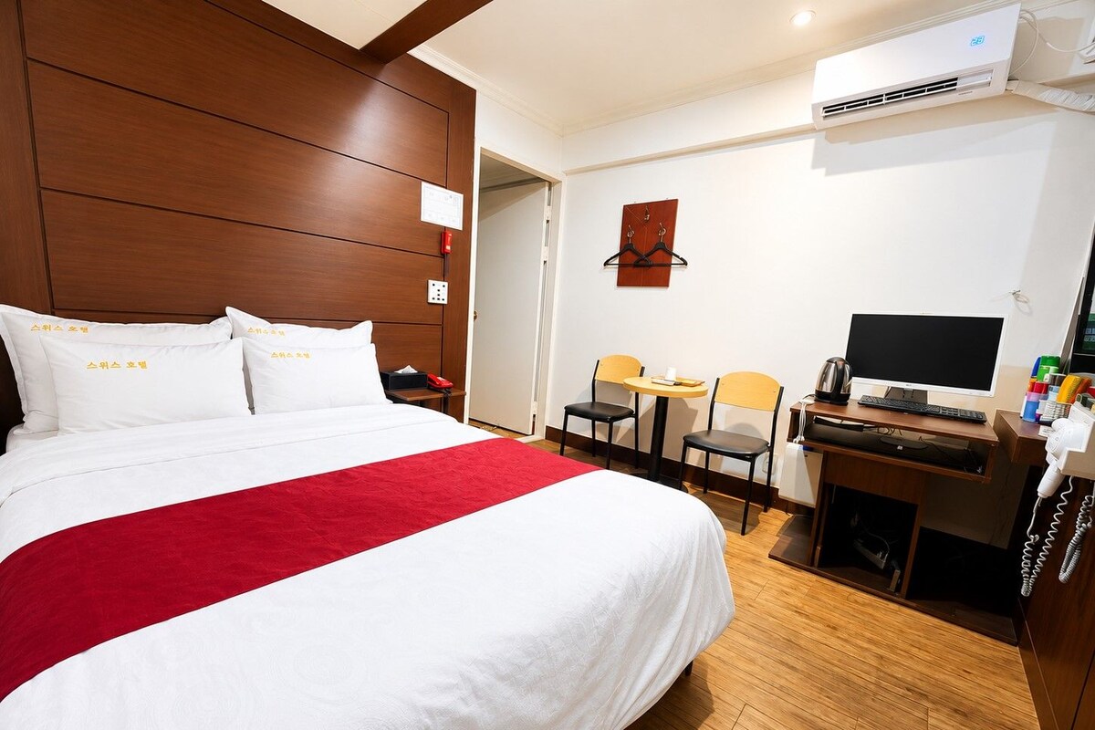 A hotel room with a large bed, a desk with a computer, two chairs, and an air conditioning unit.