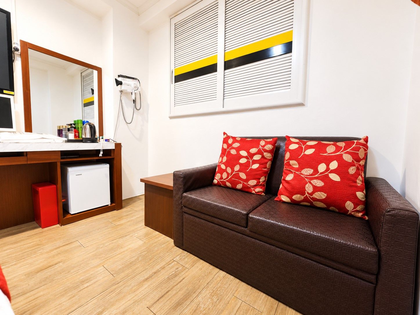 Habitación de hotel con sofá de cuero marrón, refrigerador blanco pequeño, escritorio de madera con espejo y secador de pelo incorporado.